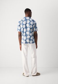 Korte mouwen, blauwe Hawaiiaanse blouse met een wit bladerenpatroon, gecombineerd met witte broek en lichtgekleurde sandalen, gezien vanuit de achterkant.