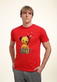 Červené bavlněné tričko s grafikou žlutého medvěda, který drží kouzelnickou hůlku, s nápisem "SOOTY" velkými písmeny pod tím.