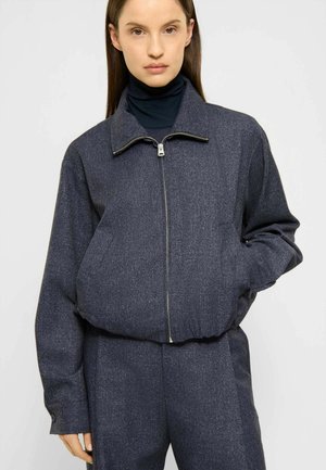 Jeune femme portant une veste zippée gris foncé sur un col roulé noir, debout avec une main dans la poche sur un fond uni.
