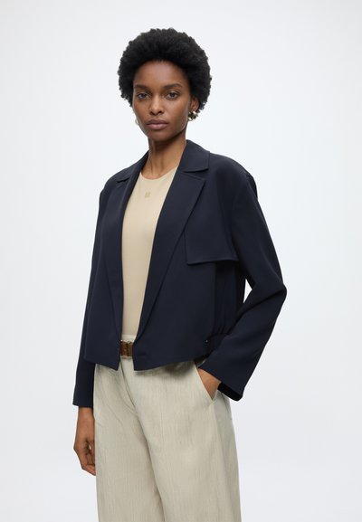 Femme aux cheveux courts et bouclés portant un blazer bleu marine raccourci, une chemise beige, un pantalon beige et une ceinture marron, debout avec une main dans la poche.