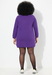 Violetti huppari-mekko, jossa on pitkät hihat, ribbaatut hihansuut ja rento istuvuus. Mekossa on suora helma, ja se on yhdistetty mustiin sukkahousuihin ja lenkkareihin.