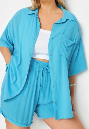 Mujer con camisa azul claro holgada abotonada y shorts a juego con cordón, sobre una camiseta blanca corta, de pie contra un fondo blanco.