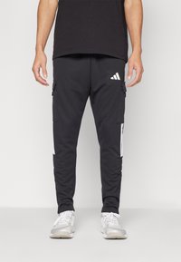 Melnas sporta bikses, kas izgatavotas no mīksta materiāla, ar kabatām, baltām sānu svītrām un Adidas logo priekšpusē.