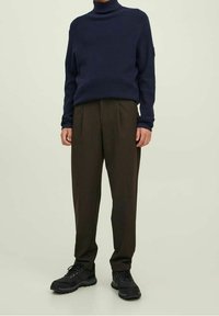 Uomo vestito con un maglione a collo alto blu navy, pantaloni marroni scuri plissettati e scarpe nere con lacci, in piedi su uno sfondo semplice.