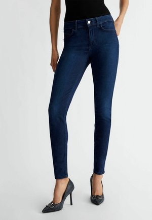 Femme portant un jean slim bleu foncé, un top noir sans manches et des talons hauts noirs pointus avec détail boucle, debout devant un fond blanc.