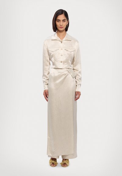 Beige satin two-piece outfit featuring a cropped button-up jacket with pockets and a long skirt with a wrapped design. Gold-accented sandals.