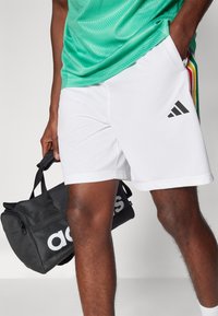 Calções atléticos brancos com um tecido texturizado, apresentando um logo Adidas preto. O modelo segura uma mala de viagem preta com uma marcação branca.