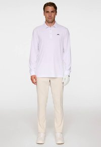 Biała koszulka polo z długim rękawem, kołnierzykiem i czarnym logo, zestawiona z beżowymi spodniami i białymi sneakersami; model nosi białą rękawiczkę.