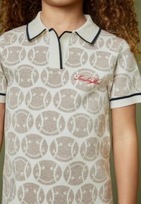 Beige ja kreem värvi polo särk, millel on korduv naeratava näo muster, kontrastne tumesinine krae ja punane tikandiga "SmileyM" tekst. Pehme tekstuur.