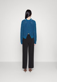 Le mannequin porte un pull en tricot bleu avec un dos découpé, associé à un pantalon noir à jambe ample et des talons avec lanières, posant devant un fond neutre.