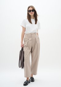 Beige wide-leg trousers with pleats, paired with a white crew neck t-shirt and black loafers, holding a brown shoulder bag, against a white background.