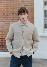 Beige textured jacket with a collared neckline, two front pockets, and button closure, paired with blue denim jeans.
