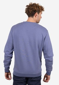 Hombre con cabello rizado que lleva una sudadera color púrpura claro y pantalones oscuros, visto desde atrás sobre un fondo blanco.