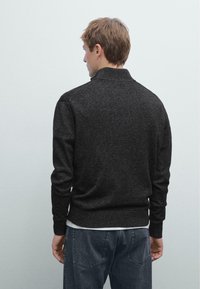 Pull en tricot gris foncé avec un col et des poignets côtelés, présentant une coupe ajustée et une texture douce. Porté par-dessus une chemise blanche.