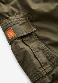 Rukáv olivovo zeleného bavlneného saka s nášivkou s nápisom "Superdry Overall", manžetou z textúrovaného materiálu a detailmi s posilneným prešívaním.
