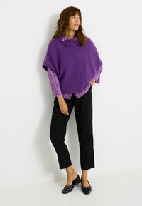 Poncho morado de punto con cuello vuelto, llevado sobre una camisa de rayas abotonada. Pantalones negros recortados y zapatos planos con lunares. Diseño simple y con textura.