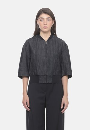Donna con capelli castani lisci che indossa una giacca corta grigio scuro con maniche corte e zip e pantaloni neri a gamba larga, in piedi su sfondo bianco.