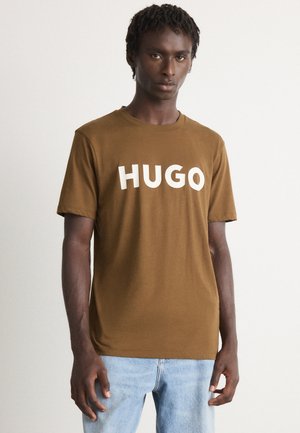 Mann mit dunkler Haut und kurzen, lockigen Haaren, der ein braunes HUGO T-Shirt und hellblaue Jeans trägt und vor einem einfarbigen, hellen Hintergrund steht.