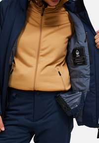 Person, der einen hellbraunen Reißverschluss-Hoodie und eine marineblaue Jacke sowie Hose trägt, hält die Jacke offen, um das schwarze Innenetikett mit weißem Text zu zeigen.
