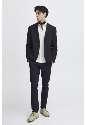 Young man wearing a black suit with a white shirt, beige shoes, and a black neck scarf, standing with hands in pockets.