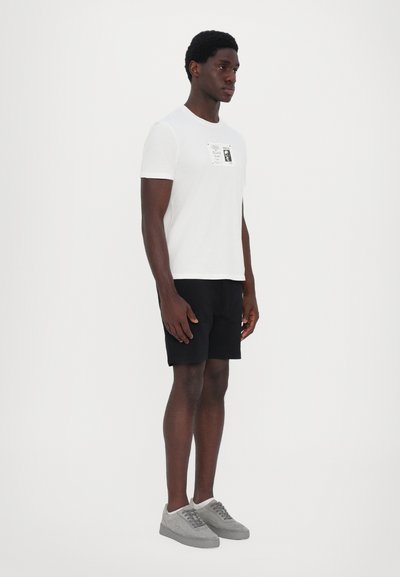 Tricou alb cu un design imprimat pe piept, asociat cu pantaloni scurți negri. Modelul poartă adidași gri, stând în fața unui fundal alb.