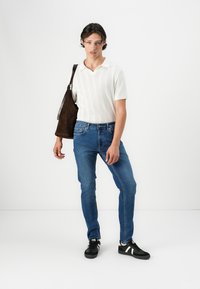 Hvit teksturert polo skjorte, slanke blå jeans, svarte sneakers med hvite striper, og en brun skulderveske. Står foran en hvit bakgrunn.