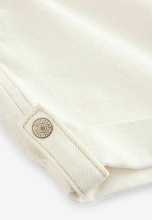 Tissu en denim blanc avec une légère texture. Présente un ourlet plié et une étiquette en forme de bouton circulaire avec "Next Jeans Since 1982" embossé.