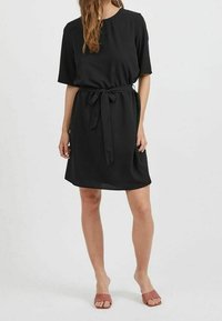 Svart tunikaklänning med korta ärmar och bälte i midjan. Slätt tyg, avslappnad passform, knälång, minimalistisk design. Bärs med rosa sandaler.