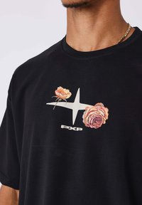 Camiseta de algodón negra con un gráfico floral que presenta rosas amarillas y rosas, un logo en forma de estrella blanca y el texto "PXP" en el centro.