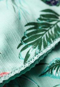 Tissu vert clair avec un motif de feuilles tropicales en vert et bleu. Présente une délicate bordure en dentelle le long d'un bord. Surface douce et texturée.