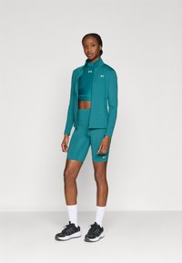 Turquoise atletische outfit bestaande uit een aansluitend jack met rits, een cropped top en een hoogwater korte broek, gecombineerd met zwarte sneakers en witte sokken.