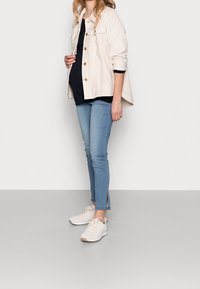 Chaqueta de mezclilla color beige claro con dos bolsillos en el pecho, camiseta negra, pantalones ajustados de mezclilla azul claro con aberturas laterales, y zapatillas claras.