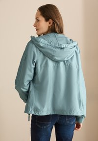Veste coupe-vent bleu clair avec capuche, présentant un intérieur texturé, des poignets élastiques et une coupe droite. Jean foncé visible en bas.