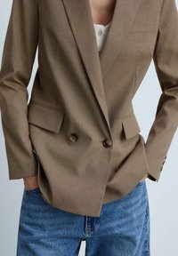 Hnědý blazer s vzorem, klopami, se dvěma knoflíky a bočními kapsami. Nosený přes světlý top a spárovaný s modrými džíny.