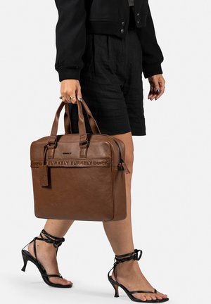 Sac fourre-tout en cuir marron avec poignées doubles, branding embossé et une étiquette sur le côté ; porté par une personne vêtue de shorts noirs et de sandales.