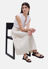 Top de lino blanco con mangas cortas, falda de lino beige y sandalias negras con acentos dorados, sentado en una silla de madera negra.