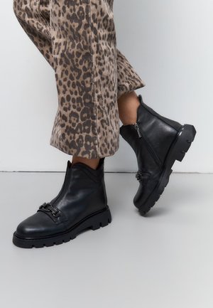 Zwarte leren enkelboots met een stevige zool, voorzien van een zijrits en decoratieve gespsluiting. Draagt men met een broek met luipaardprint.