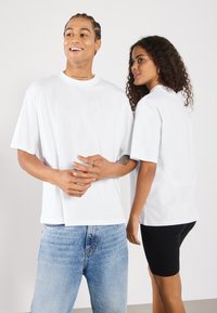 Biele oversized tričká z jemnej bavlny s krátkymi rukávmi a okrúhlym výstrihom, nosené s modrými riflami a čiernymi šortkami.