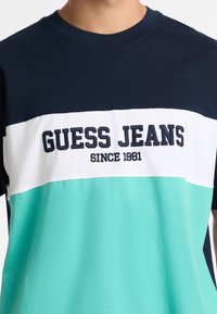 Jūras, balta un piparmētru zaļā krāsā bloku krekliņš ar apaļu kaklu. Izceļas ar "GUESS JEANS" logo un "SINCE 1981" jūras krāsā. Mīksta auduma.
