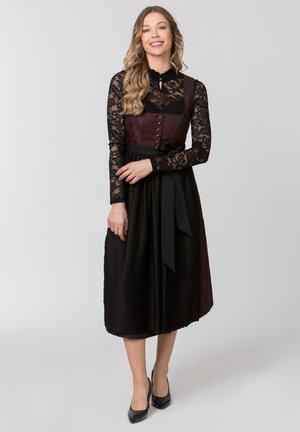 Junge Frau lächelnd, trägt ein schwarzes Spitzenoberteil und ein dunkelburgunder Dirndlkleid mit schwarzer Schürze und schwarzen High Heels, steht vor einem einfarbigen Hintergrund.