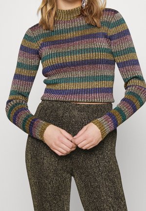 Femme portant un pull rayé métallique côtelé à manches longues et un pantalon noir pailleté taille haute, mains jointes devant.