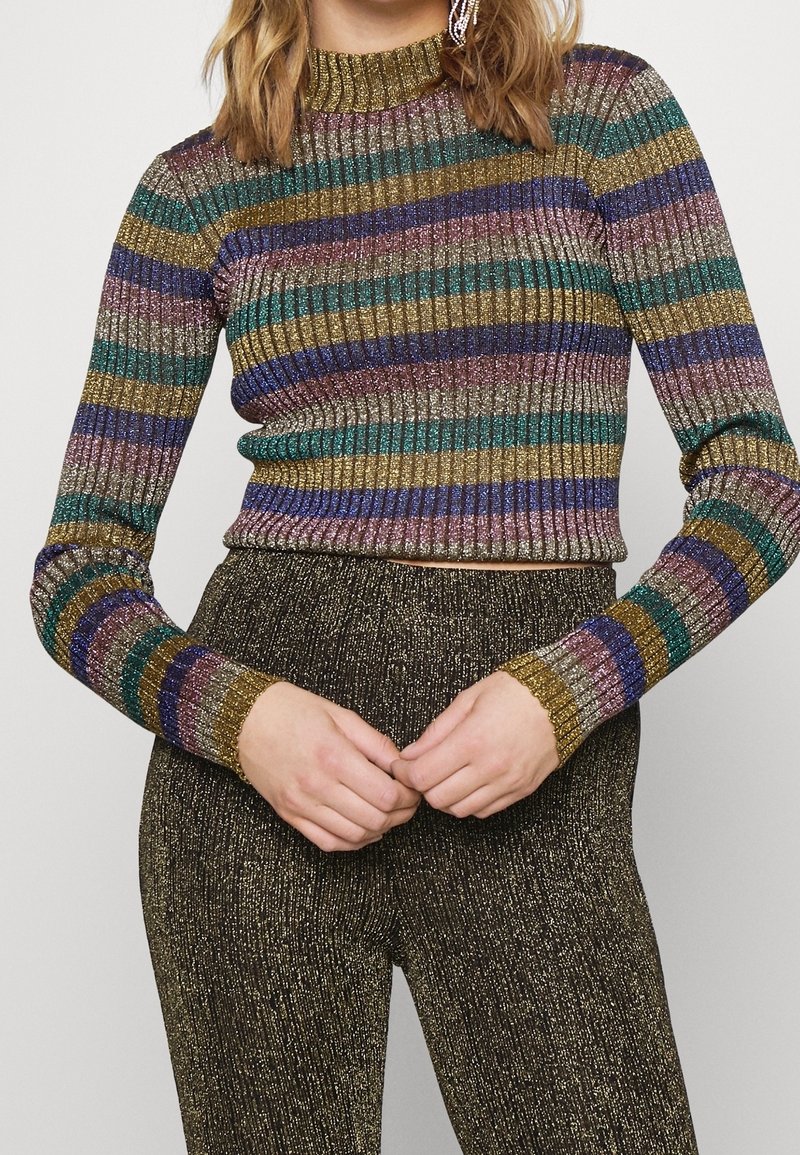 Femme portant un pull rayé métallique côtelé à manches longues et un pantalon noir pailleté taille haute, mains jointes devant.
