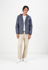 Un jeune homme se tient debout portant une veste zippée grise, un t-shirt blanc, un pantalon beige et des baskets blanches sur un fond blanc uni.