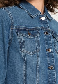 Giacca di jeans blu chiaro con chiusura a bottoni, due tasche sul petto e finiture in argento. Presenta un colletto classico e cuciture a contrasto.