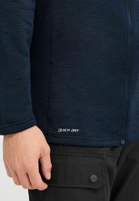 Dunkelblaue, schnell trocknende Jacke mit strukturiertem Oberflächenmaterial. Verfügt über einen Reißverschluss und den kontrastierenden Schriftzug "QUICK DRY" nahe dem Saum. Schwarze Hose mit Taschen-Detail.