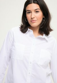Camicia bianca con bottoni e maniche lunghe, con due tasche sul petto, colletto classico e una texture di cotone liscia.