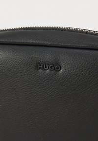 Pochette nera in pelle con finitura testurizzata, dotata di una cerniera in argento e logo "HUGO" in rilievo sul fronte. Forma compatta e rettangolare.