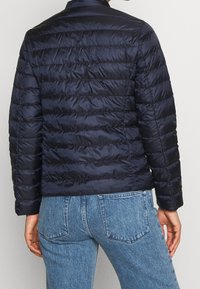 Person wearing a navy blue quilted jacket and blue denim jeans, shown from the back against a plain background.