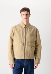 Beige bomullsjakke med glidelås, spiss krage og to frontlommer. Har en teksturert stoff og en logoklapp på venstre lomme.