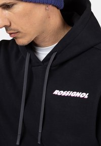 Felpa nera con coulisse grigia presenta un logo "ROSSIGNOL" bianco e rosa nella zona del petto. Il materiale sembra morbido con una texture liscia.